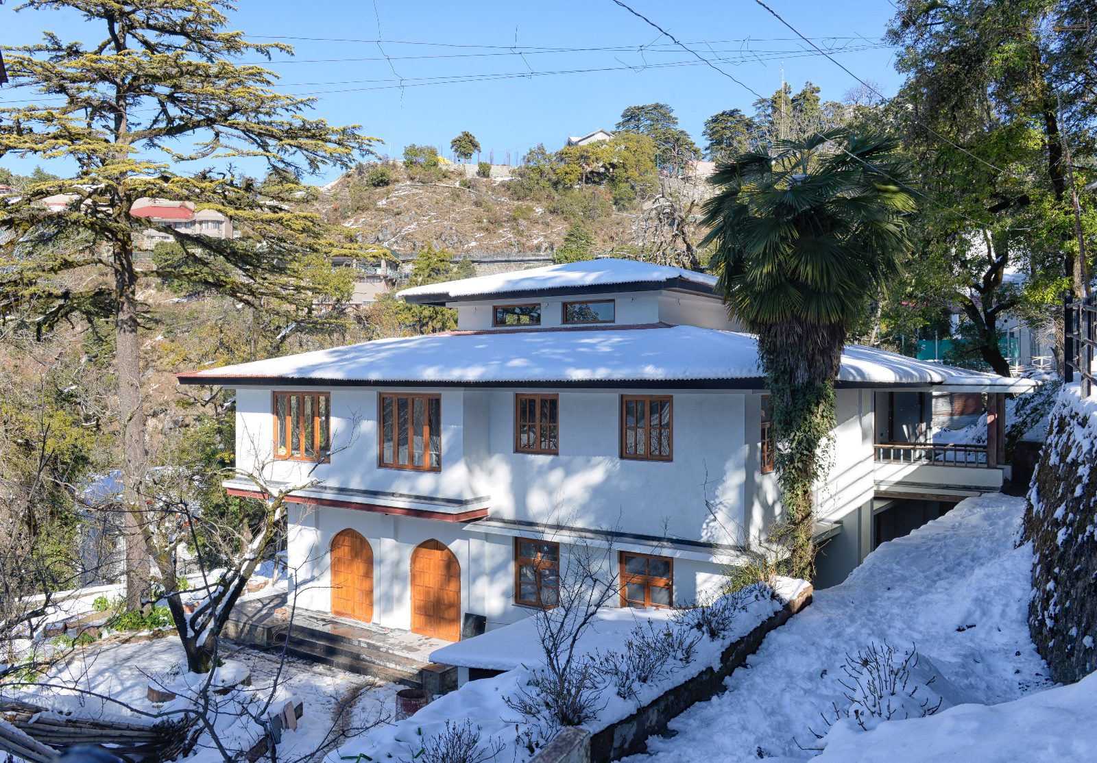 White two-storey hillside house with snow-covered tiered roofs, timber windows and arched wooden doors, among cedar trees.