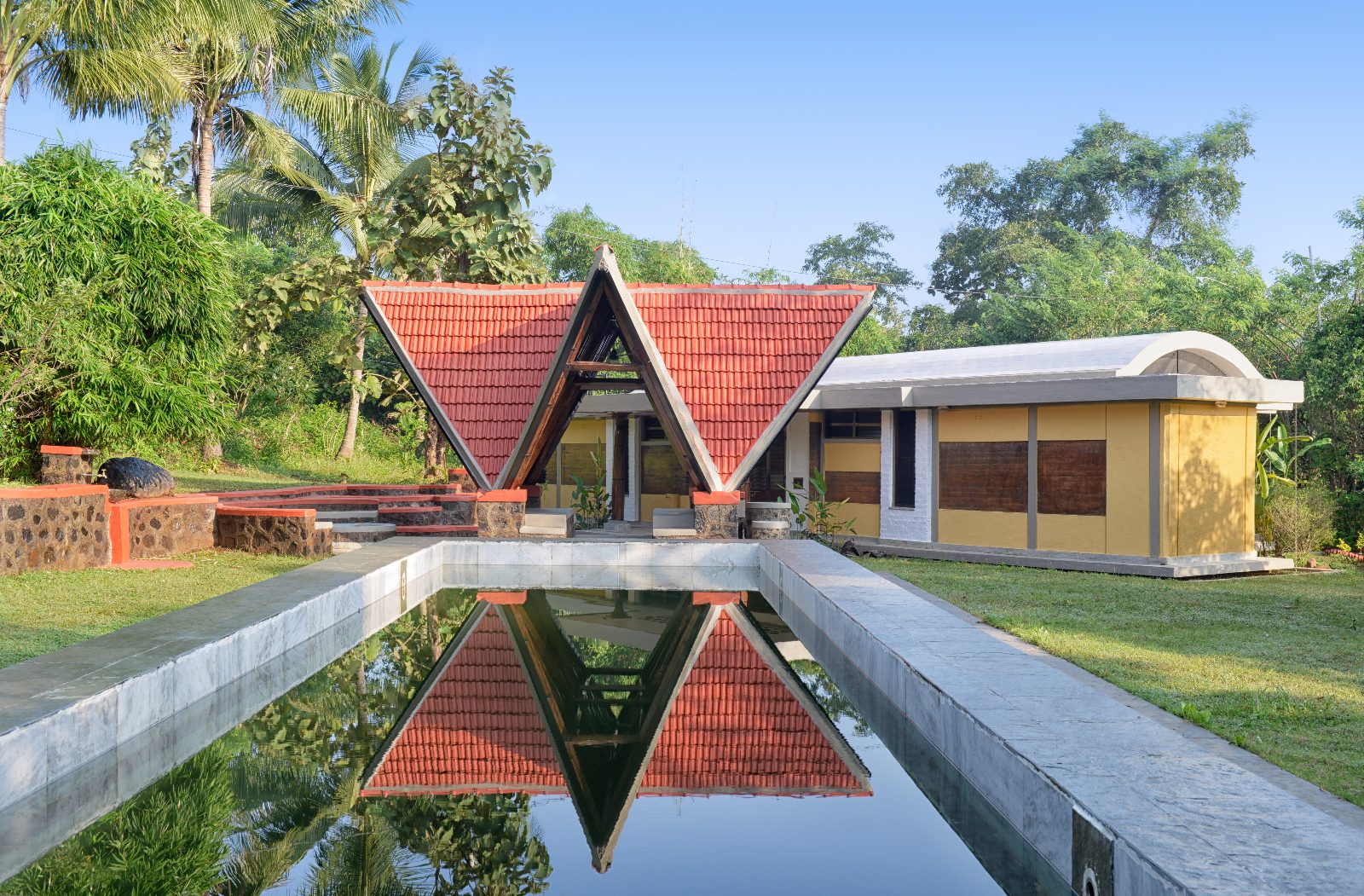 Farmhouse pavilion with a terracotta tiled A-frame roof reflected in a long stone pool, with an ochre vaulted wing and palm trees.